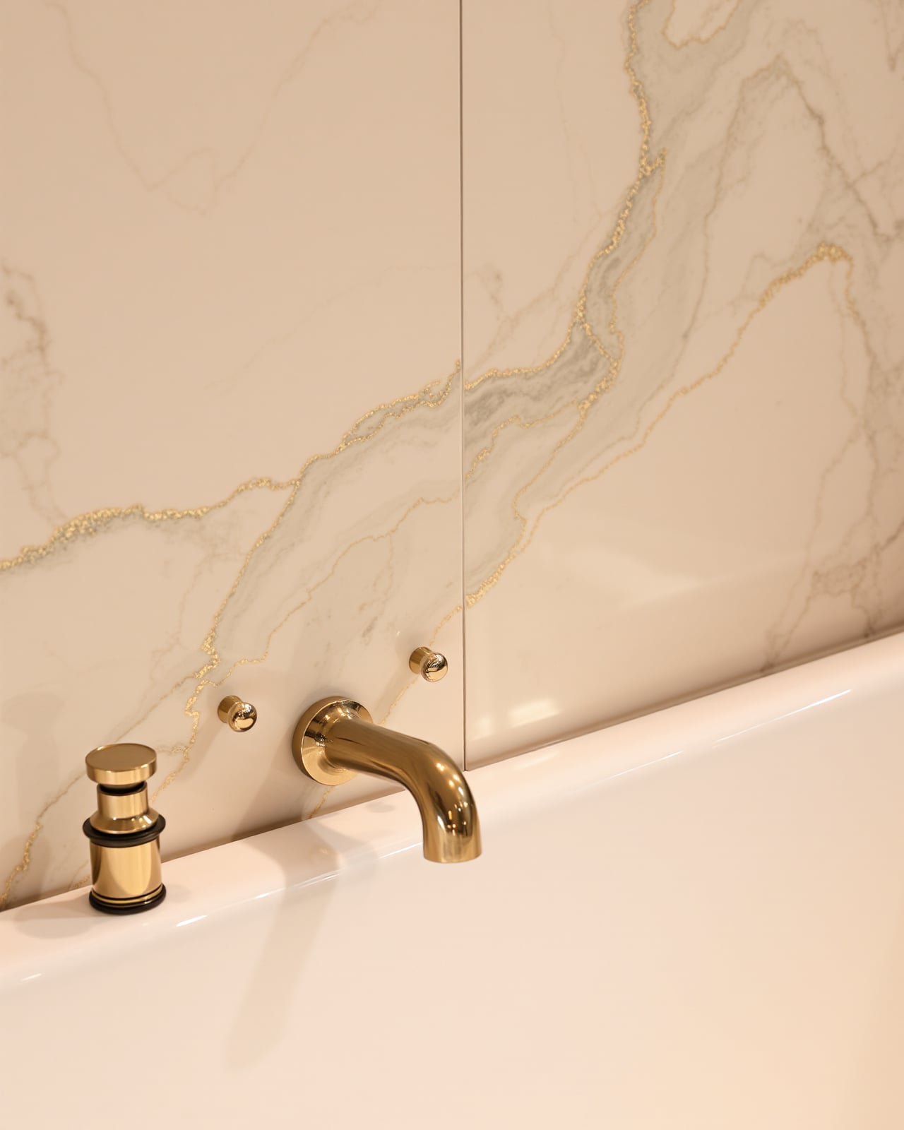A polished brass floor-mounted tub filler and lever handles against a wall of book-matched cream marble, its grey and gold veining mirrored across the seam.
